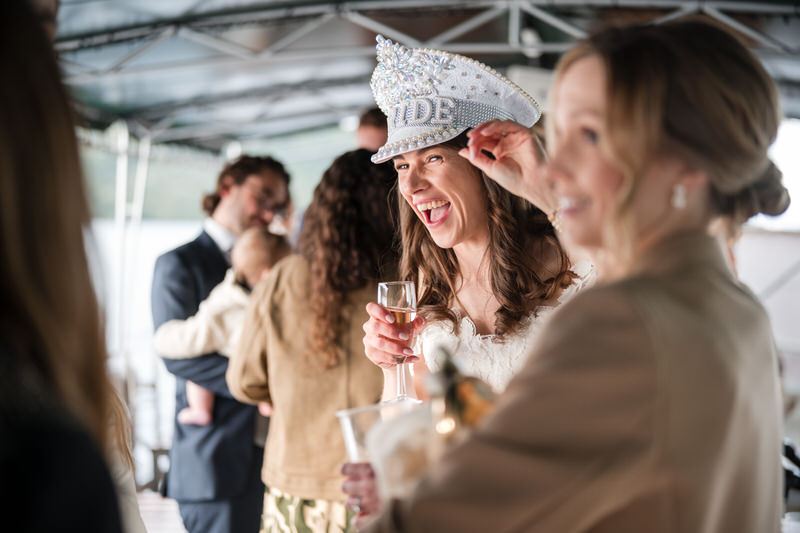 Bride wearing a hen party hat laughing with champagne during wedding reception at Inn on the Lake