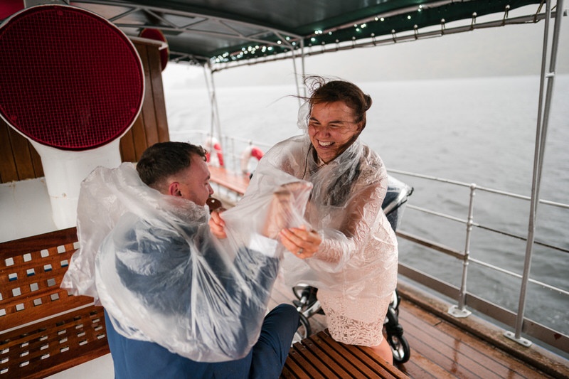 Wedding guests laughing in rain ponchos aboard a boat on Ullswater