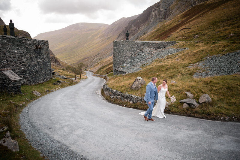 Lake District mountain road wedding couple portrait Wedding couple walking along a dramatic mountain road in the Lake District with hills rising around them