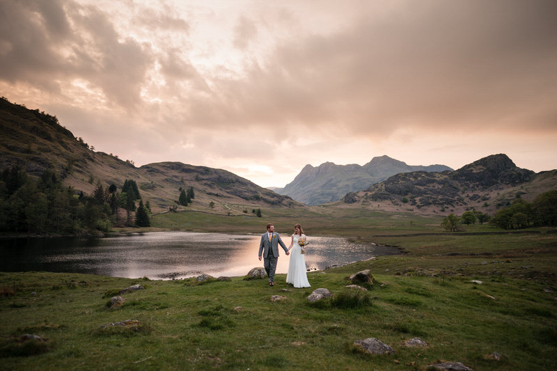 Wedding couple walking beside a Lake District lake with mountains and a soft sunset sky