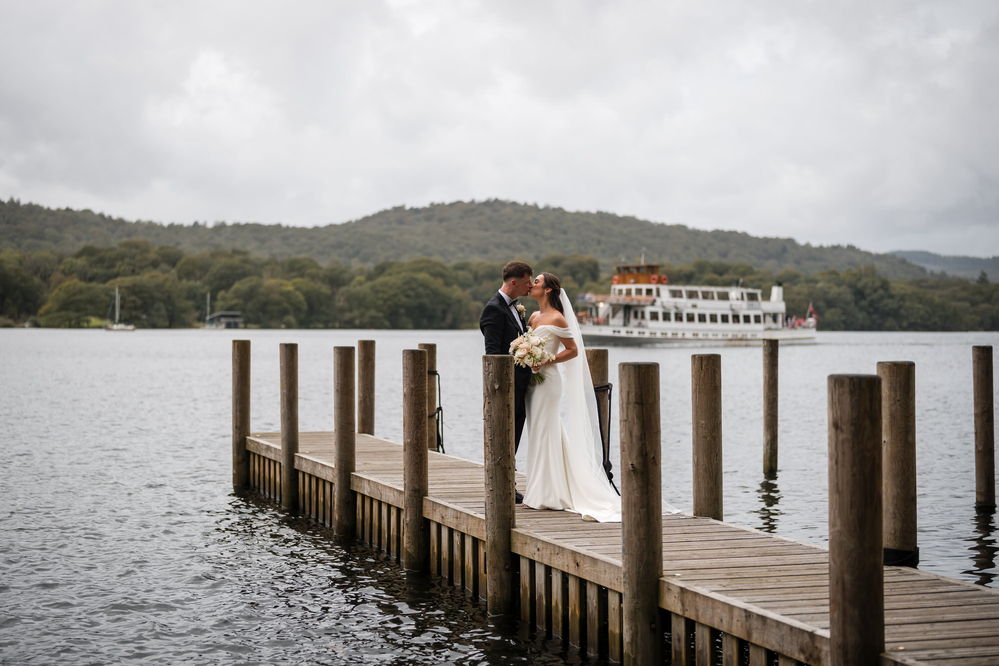 Dan and Ellie kissing on the jetty with a boat passing on Windermere